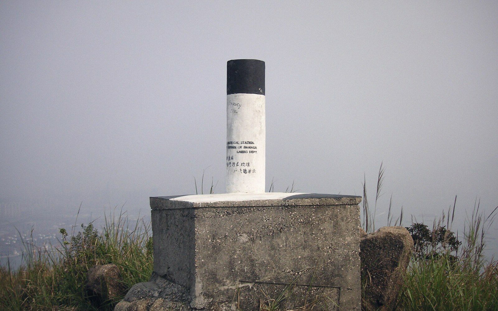 A concrete trigonometrical station pillar sits atop a hazy, grassy hilltop overlooking the landscape below
