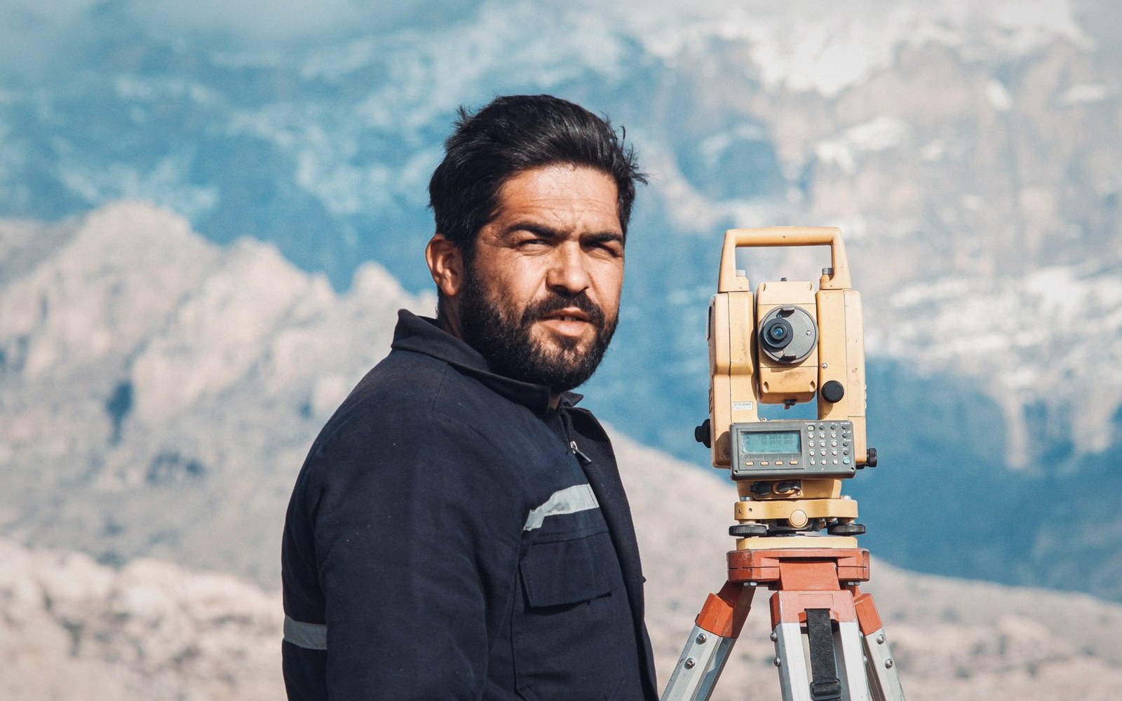 Surveyor stands beside a total station on a tripod against mountain terrain